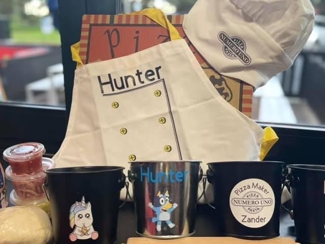An embroidered Numero Uno Pizza chef hat beside a personalised Pizza Maker metal bucket and a named child's apron on a windowsill at Numero Uno.