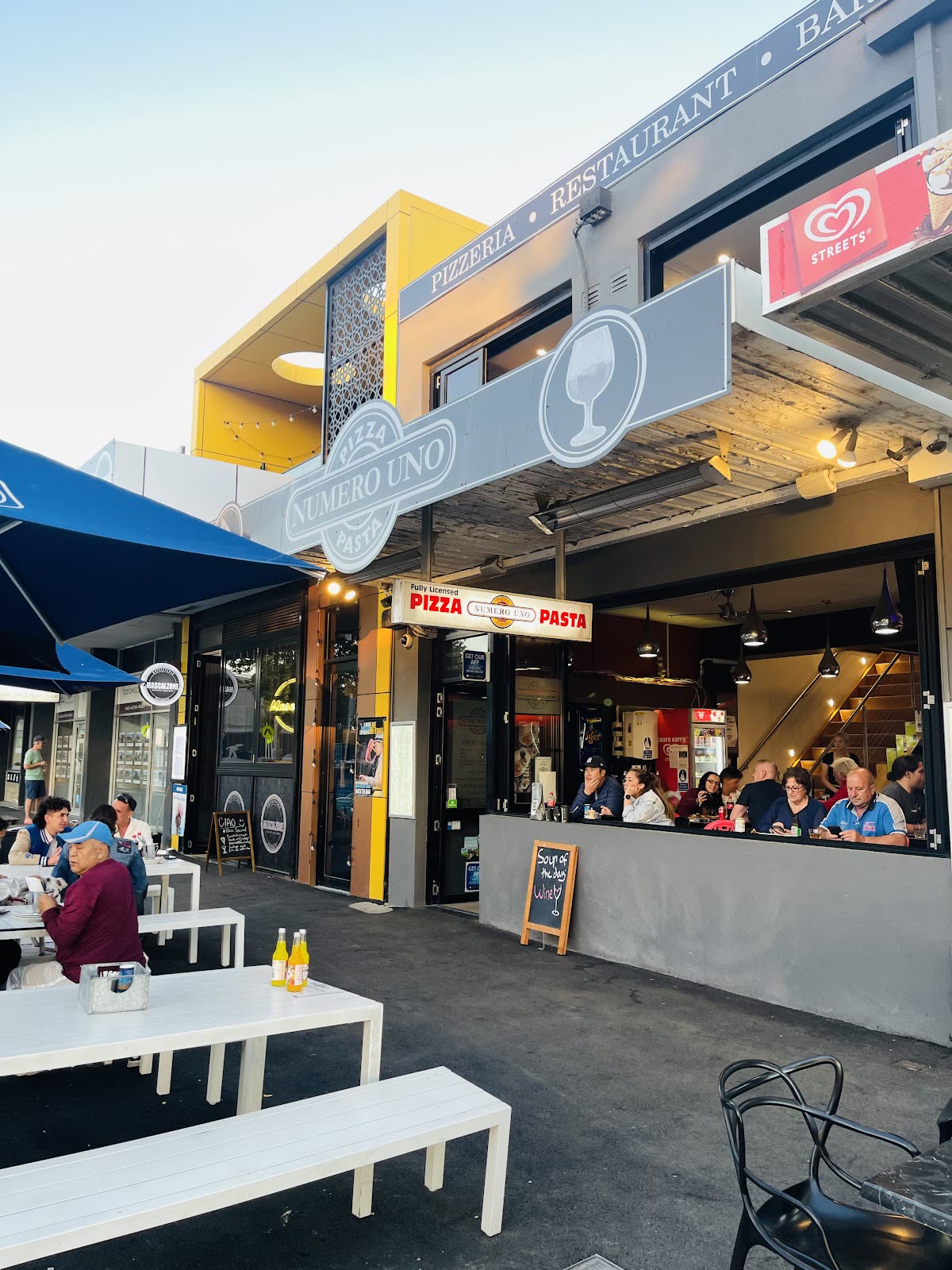 The Numero Uno Pizza and Pasta shopfront at 20 Pier Street Altona: grey oval sign with a wine glass, red Fully Licensed Pizza Pasta fascia, mustard yellow upper wall and white footpath tables.